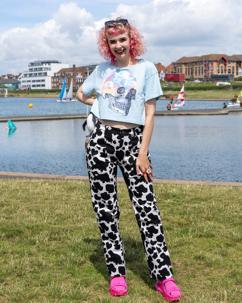 A model stands outdoors by a lakeside, smiling in a pair of Run & Fly Black & White Cow Print Straight Leg Trousers. They pair the bold monochrome pattern with a light blue cropped skull graphic T-shirt and bright pink sandals. The trousers are part of Run & Fly's ethical slow fashion range, perfect for casual, alternative styling. Sailboats and colourful buildings are visible in the background.