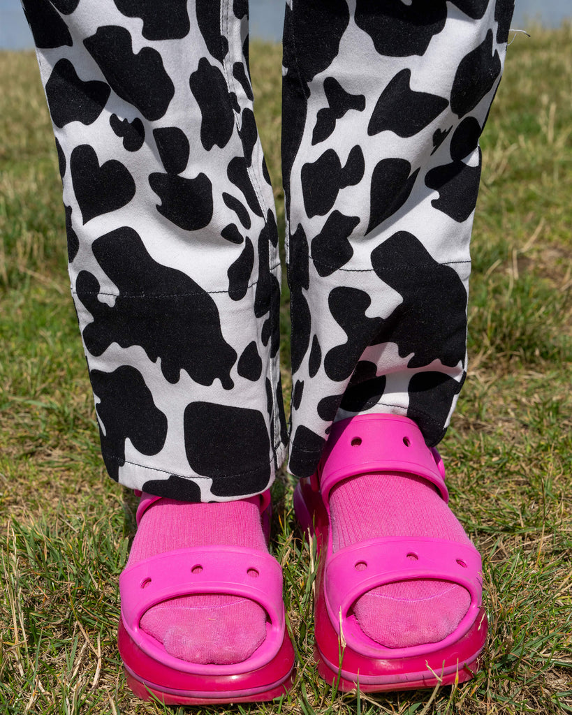 A close-up view of the lower half of a model wearing Run & Fly's Black & White Cow Print Straight Leg Trousers. The bold black and white cow pattern is paired with bright pink sandals and matching socks. The trousers feature a straight-leg cut and are designed by Run & Fly, a small ethical slow fashion brand known for quirky and playful prints.