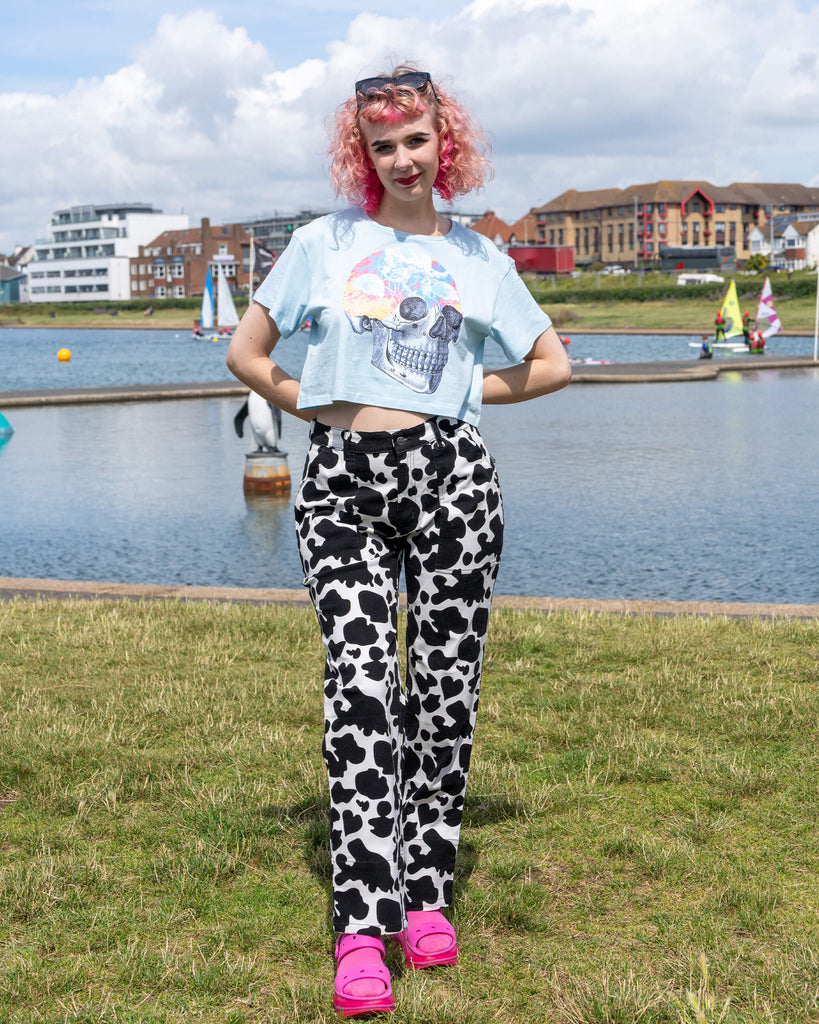 The model stands confidently by a lake, hands on hips, wearing Run & Fly Black & White Cow Print Straight Leg Trousers. The playful cow print is complemented by a light blue cropped top with a vibrant skull design and bright pink sandals. The slow fashion brand Run & Fly offers fun, retro-inspired clothing with an ethical production ethos.