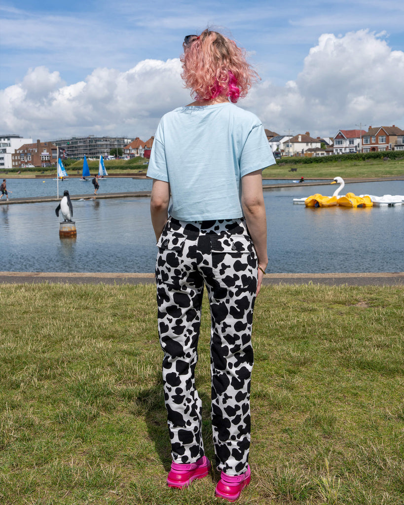 The model faces away from the camera, showing the back view of the Run & Fly Black & White Cow Print Straight Leg Trousers. The playful cow print runs throughout the design, paired with a light blue cropped top and bright pink sandals. Run & Fly is a small ethical slow fashion company known for its bold, retro-inspired clothing.