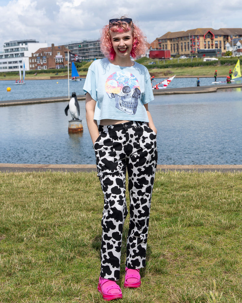 The model stands outdoors in the sun, wearing Run & Fly Black & White Cow Print Straight Leg Trousers. The bold monochrome pattern is paired with a light blue cropped skull T-shirt and pink sandals. The trousers are part of Run & Fly’s ethical slow fashion collection, offering a playful, alternative style.