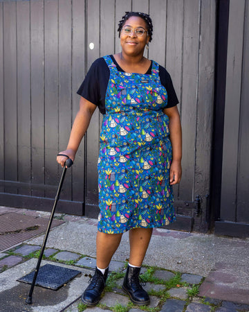 Han, a black femme model with silver glasses with a chain, is stood outside in Hove in a mews wearing Run & Fly x Katie Abey Crystal Critters Stretch Twill Long Pinafore Dress with a black t shirt underneath and black boots. The blue pinafore dress has an all over print of critters and crystals illustrated by Katie Abey. Han is smiling toward the camera with one hand in the pinafore pocket and one leg in front of the other. She is leaning on her black walking aid. 