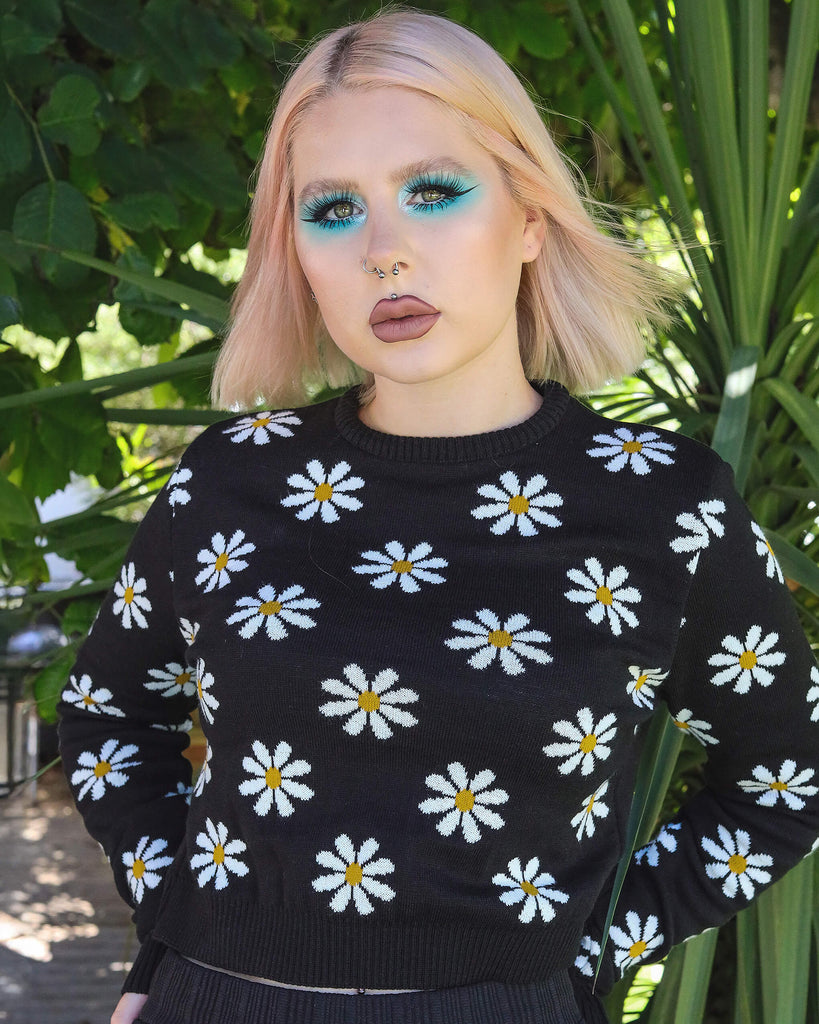 Model is stood in a green garden area wearing the daisy chain black cropped jumper with black trousers. They are facing the camera with both hands on their hips. Photo is cropped from the hips up.