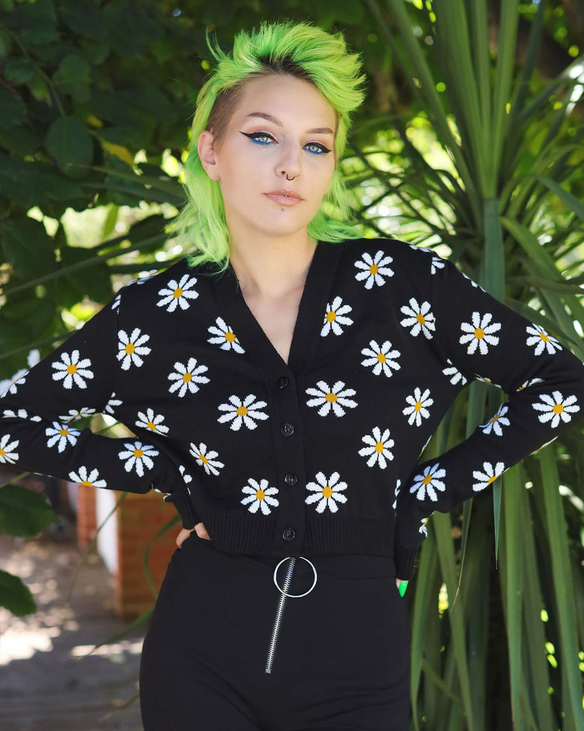Green haired model is stood nearby a tree wearing the daisy chain black cropped cardigan and black trousers. They are facing the camera smising with both hands on their hips. Photo is cropped from the knees up.