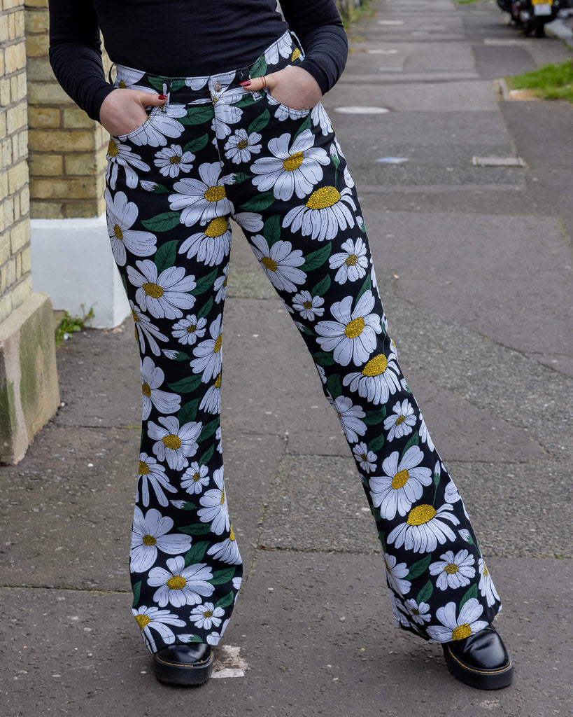  a person wearing Run & Fly daisy flare trousers outdoors in Hove. These trousers feature a white daisy print with green leaves and yellow centres on a black background. With a high-waisted fit and flared legs, they offer a retro-inspired style. The model has their hands in the pockets of the flares