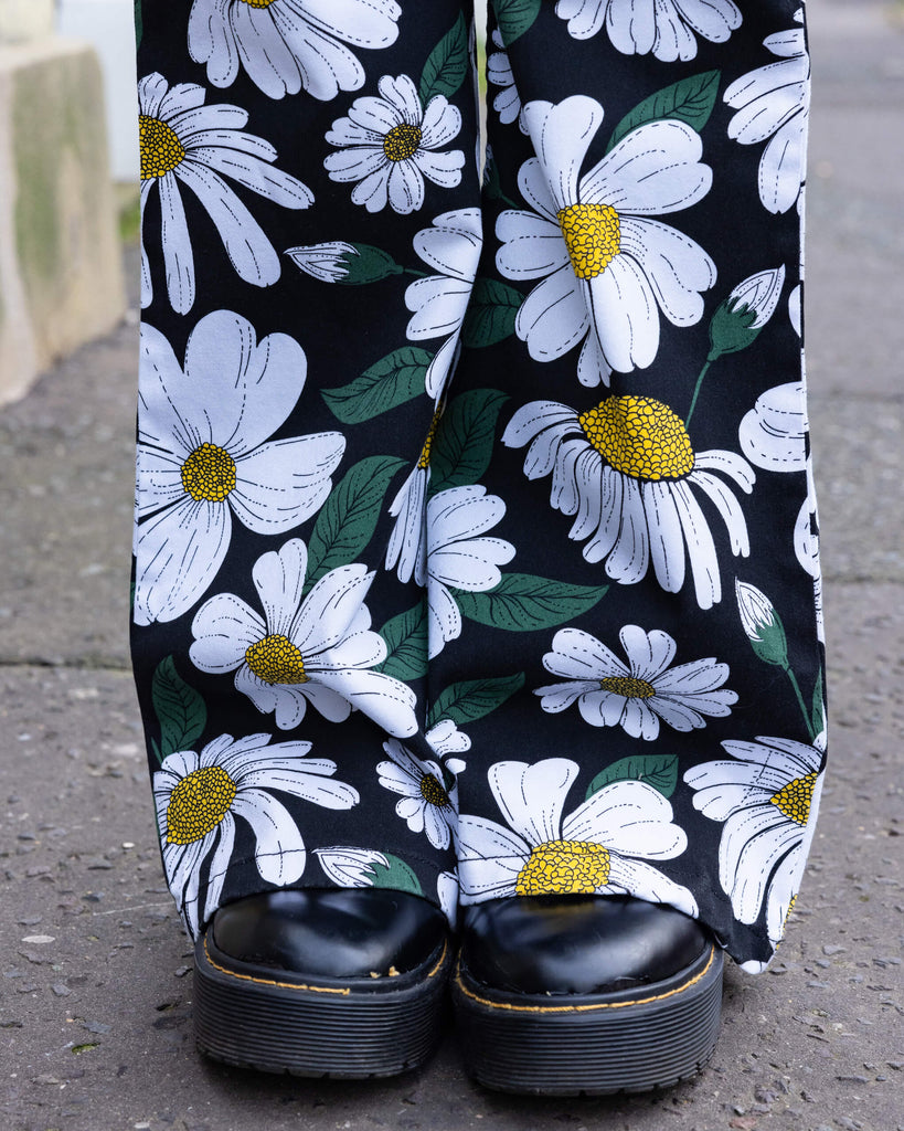 Close-up of the flared bottoms of Run & Fly daisy flare trousers with black boots peeping out the bottom. Featuring a bold white daisy print with green leaves and yellow centres on a black background. The photo is taken outdoors in Hove