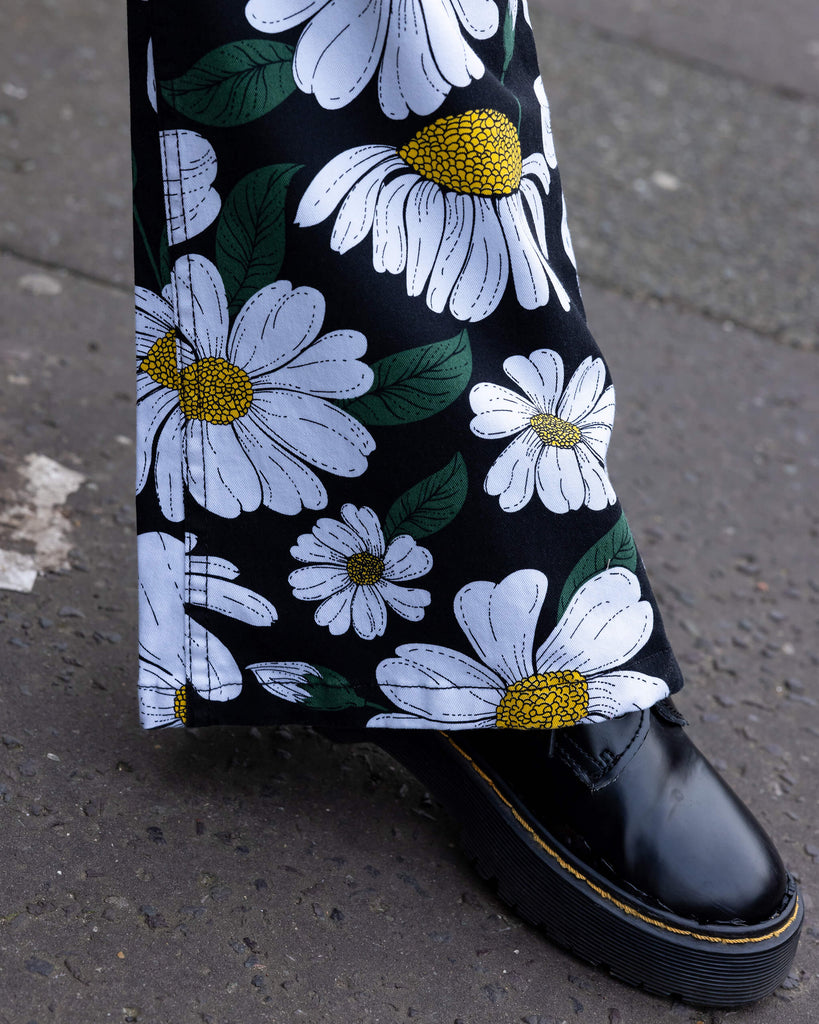 Close-up of the flared bottoms of Run & Fly daisy flare trousers with black boots peeping out the bottom. Featuring a bold white daisy print with green leaves and yellow centres on a black background. The photo is taken outdoors in Hove