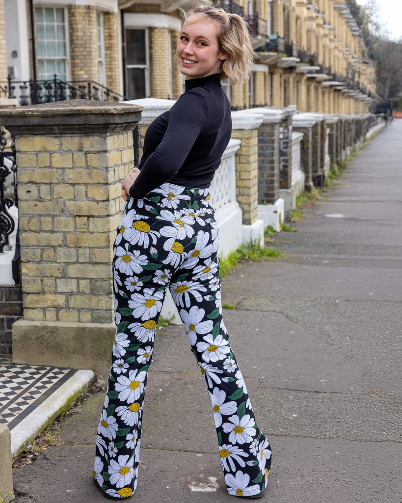 Blonde femme model stood outdoors in Hove wearing Run & Fly daisy flare trousers outdoors in Hove. These trousers feature a white daisy print with green leaves and yellow centres on a black background. With a high-waisted fit and flared legs, they offer a retro-inspired style. The model is looking over her shoulder smiling at the camera with her hand on her hip