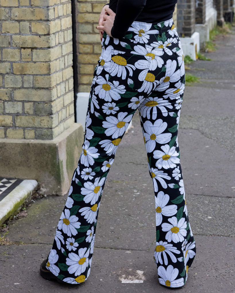 a person wearing Run & Fly daisy flare trousers outdoors in Hove. These trousers feature a white daisy print with green leaves and yellow centres on a black background. With a high-waisted fit and flared legs, they offer a retro-inspired style.