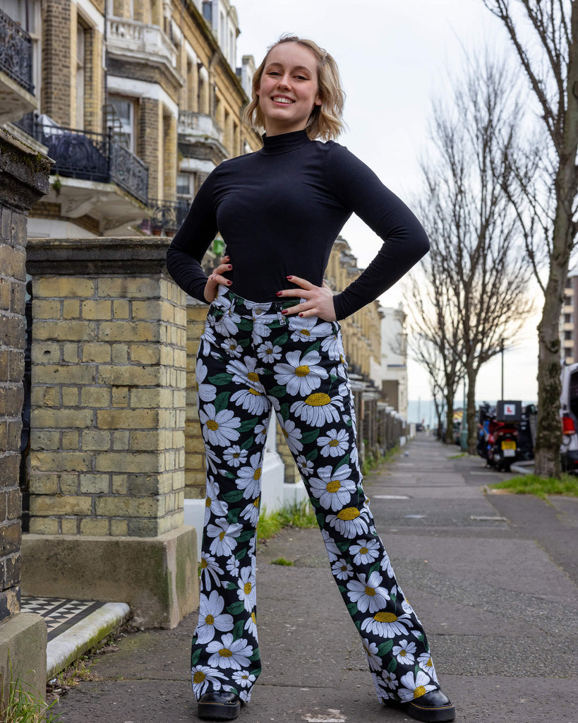 Blonde femme model stood outdoors in Hove wearing Run & Fly daisy flare trousers outdoors in Hove. These trousers feature a white daisy print with green leaves and yellow centres on a black background. With a high-waisted fit and flared legs, they offer a retro-inspired style. The model is posing and smiling at the camera with her hands on her hips