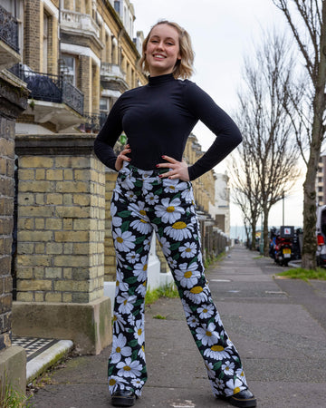 Blonde femme model stood outdoors in Hove wearing Run & Fly daisy flare trousers outdoors in Hove. These trousers feature a white daisy print with green leaves and yellow centres on a black background. With a high-waisted fit and flared legs, they offer a retro-inspired style. The model is posing and smiling at the camera with her hands on her hips