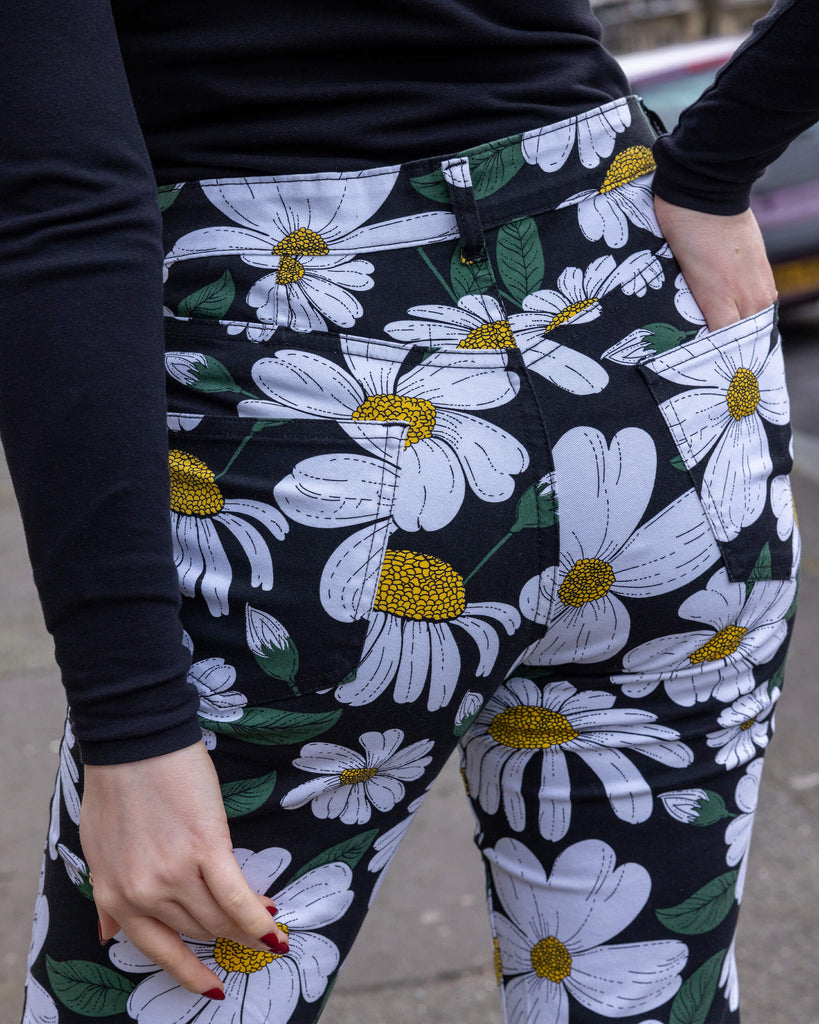 Close-up of the back pocket area of Run & Fly daisy flare trousers. Featuring a bold white daisy print with green leaves and yellow centres on a black background. The photo is taken outdoors in Hove. The model's hand is resting in the back pocket of the flares