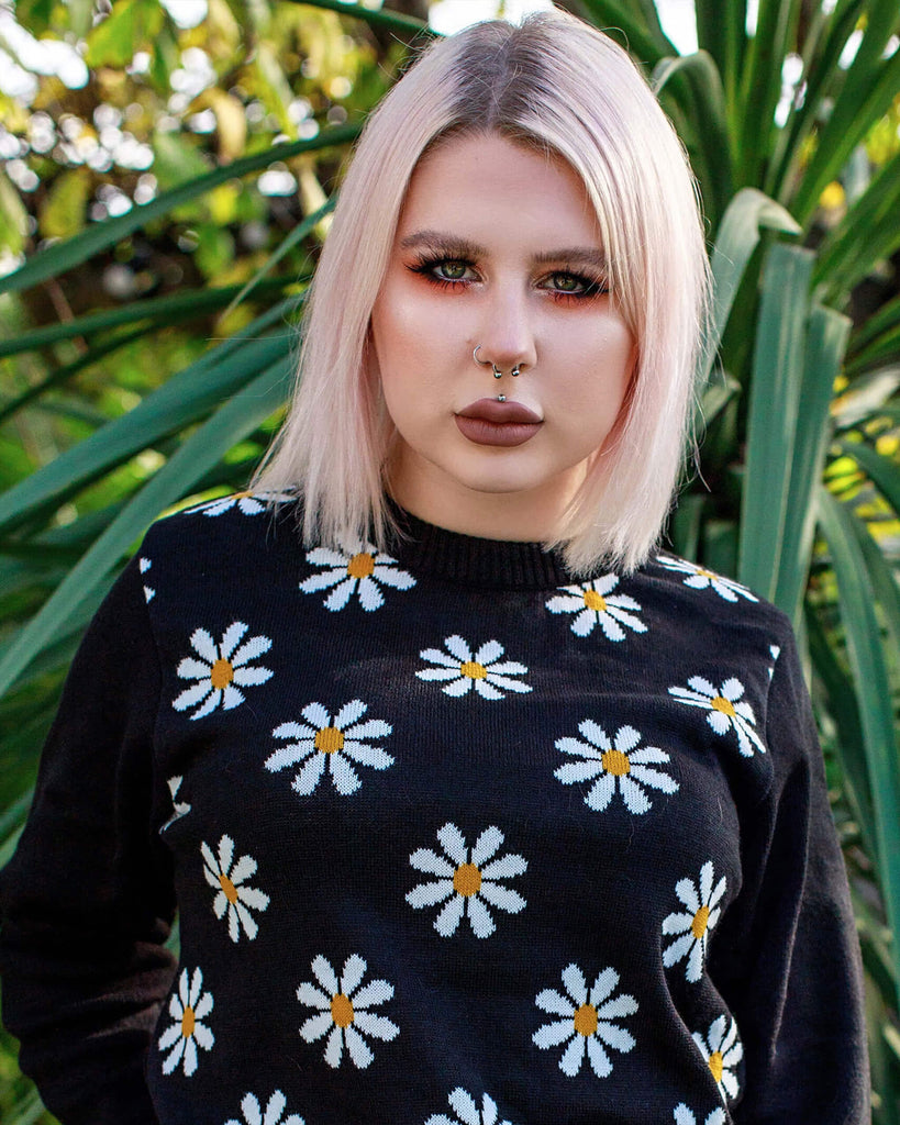 Florence is wearing the Daisy Chain Black Jumper and has a blonde sharp bob and orange eye makeup, she is stood facing forward outside in a green garden. The black jumper has plain black arms and back with repeating white daisies across the front body.