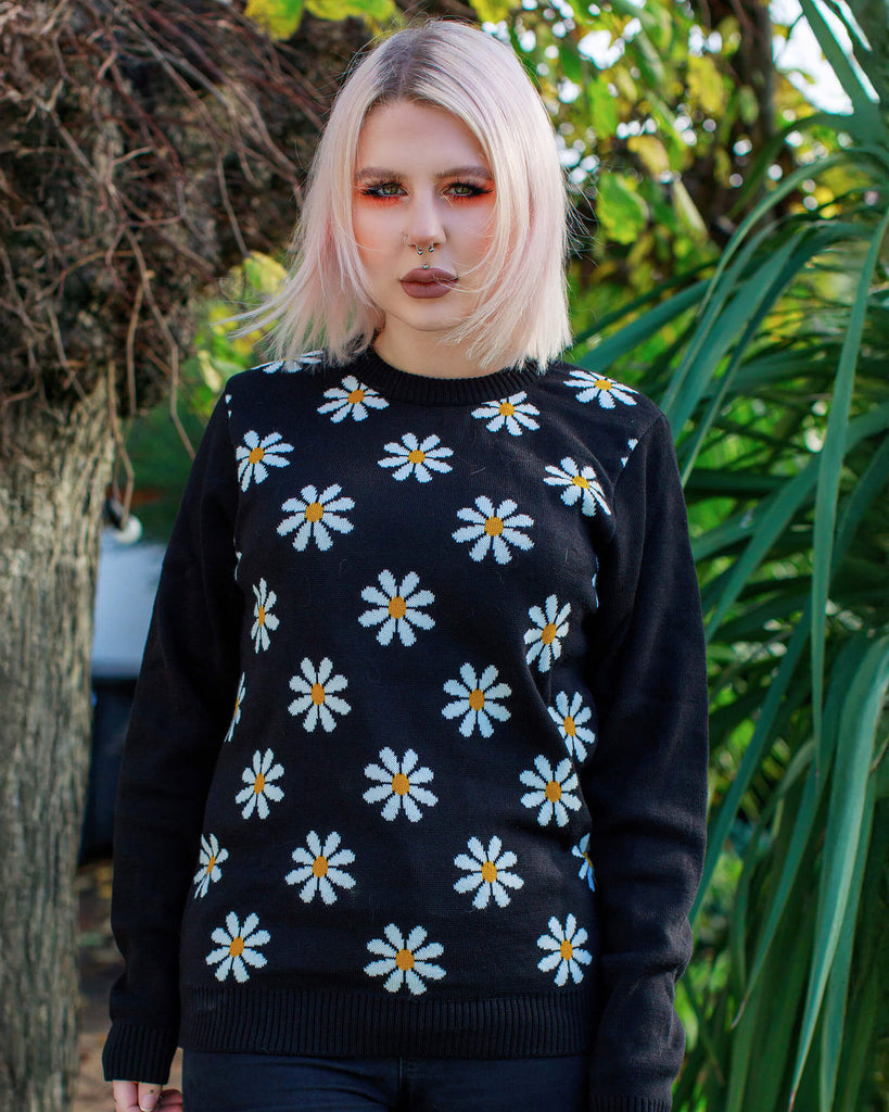 Florence is wearing the Daisy Chain Black Jumper and has a blonde sharp bob and orange eye makeup, she is stood facing forward outside in a green garden. The black jumper has plain black arms and back with repeating white daisies across the front body.