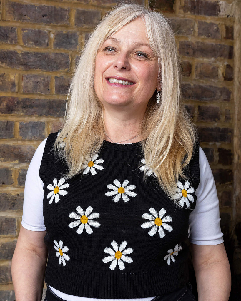 karina a white woman in her 50's has blonde hair and is wearing a daisy knitted tank top and jeans with a white tee against a brick wall