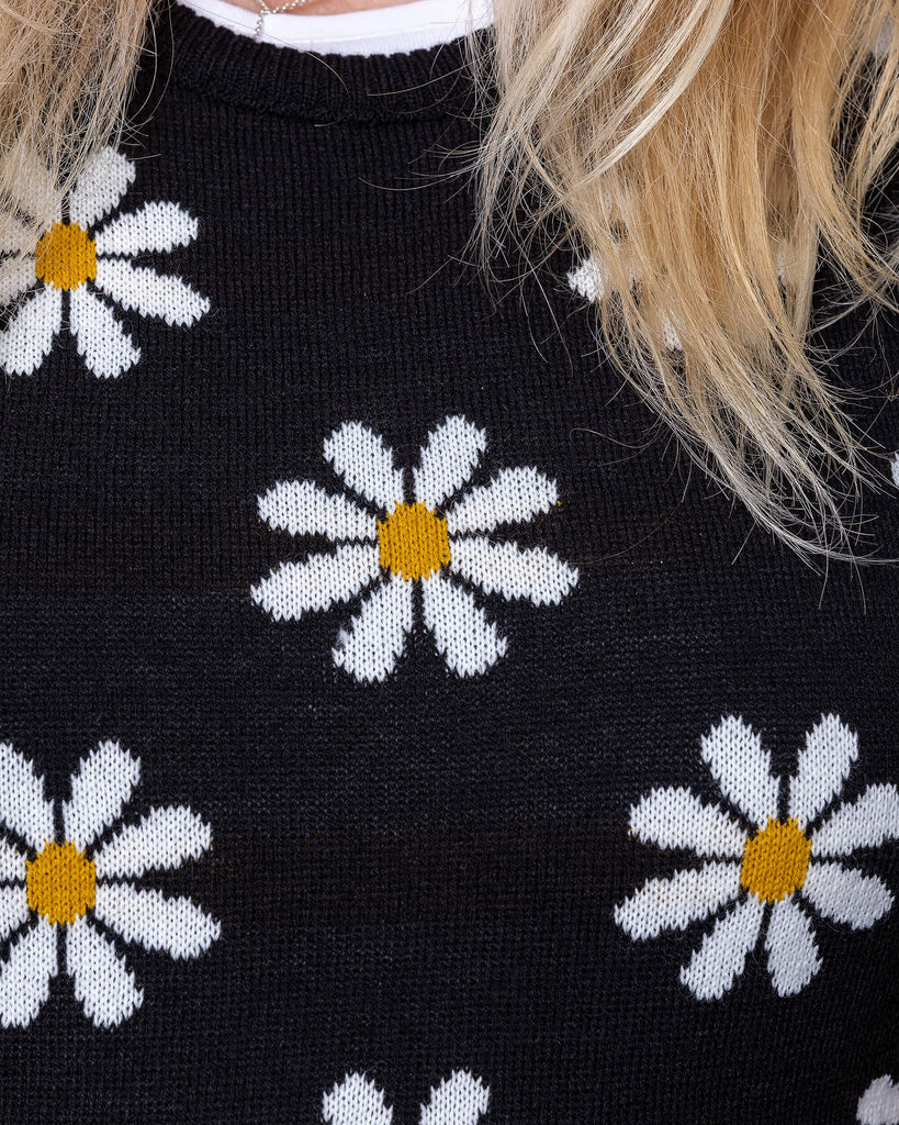 Close up of karina a white woman in her 50's has blonde hair and is wearing a daisy knitted tank top and jeans with a white tee against a brick wall