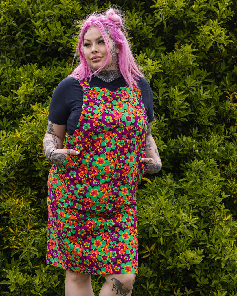 Zoe is stood in a garden area wearing the 70's dark floral print stretch corduroy long pinafore dress with a short sleeve black tshirt and black boots. She has long pink hair in half up space buns, she's facing forward posing with both hands in the pinafore pockets whilst looking off to the left.