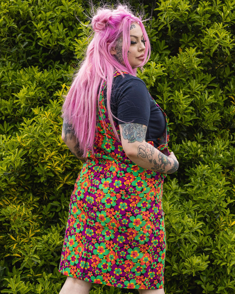 Zoe is stood in a garden area wearing the 70's dark floral print stretch corduroy long pinafore dress with a short sleeve black tshirt and black boots. She has long pink hair in half up space buns, she's facing away from the camera posing with both hands in the pinafore pockets whilst looking down over her right shoulder.