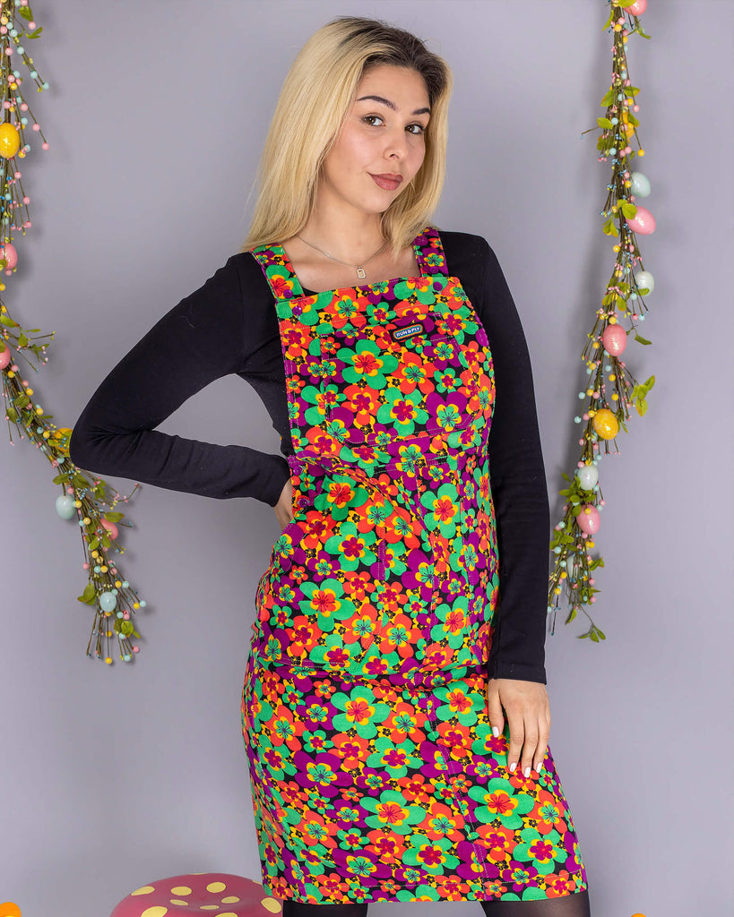 Shannon is stood in front of a grey studio background with various floral and easter decorations whilst wearing the 70's dark floral print stretch corduroy long pinafore dress with a long sleeve black tshirt, black tights and black boots. They are facing forward smiling with one hand on their hip.