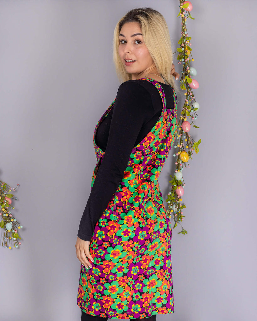 Shannon is stood in front of a grey studio background with various floral and easter decorations whilst wearing the 70's dark floral print stretch corduroy long pinafore dress with a long sleeve black tshirt, black tights and black boots. They are facing away from the camera looking over their right shoulder smiling.