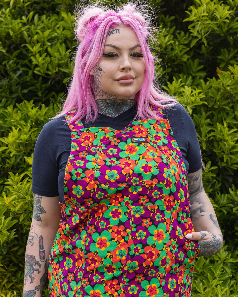 Zoe is stood in a garden area wearing the 70's dark floral print stretch corduroy long pinafore dress with a short sleeve black tshirt and black boots. She has long pink hair in half up space buns, she's facing forward posing with both hands resting by her side and smiling to camera. Photo is cropped from the knees up.