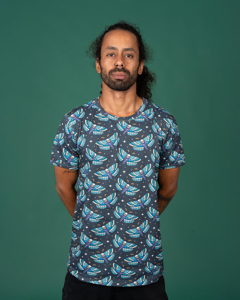 Model standing against a green background wearing a dark grey T-shirt by Run & Fly, featuring blue and teal death head moths with skulls. The moths are repeated across the shirt. Ethical slow fashion focusing on alternative, gothic-inspired designs.