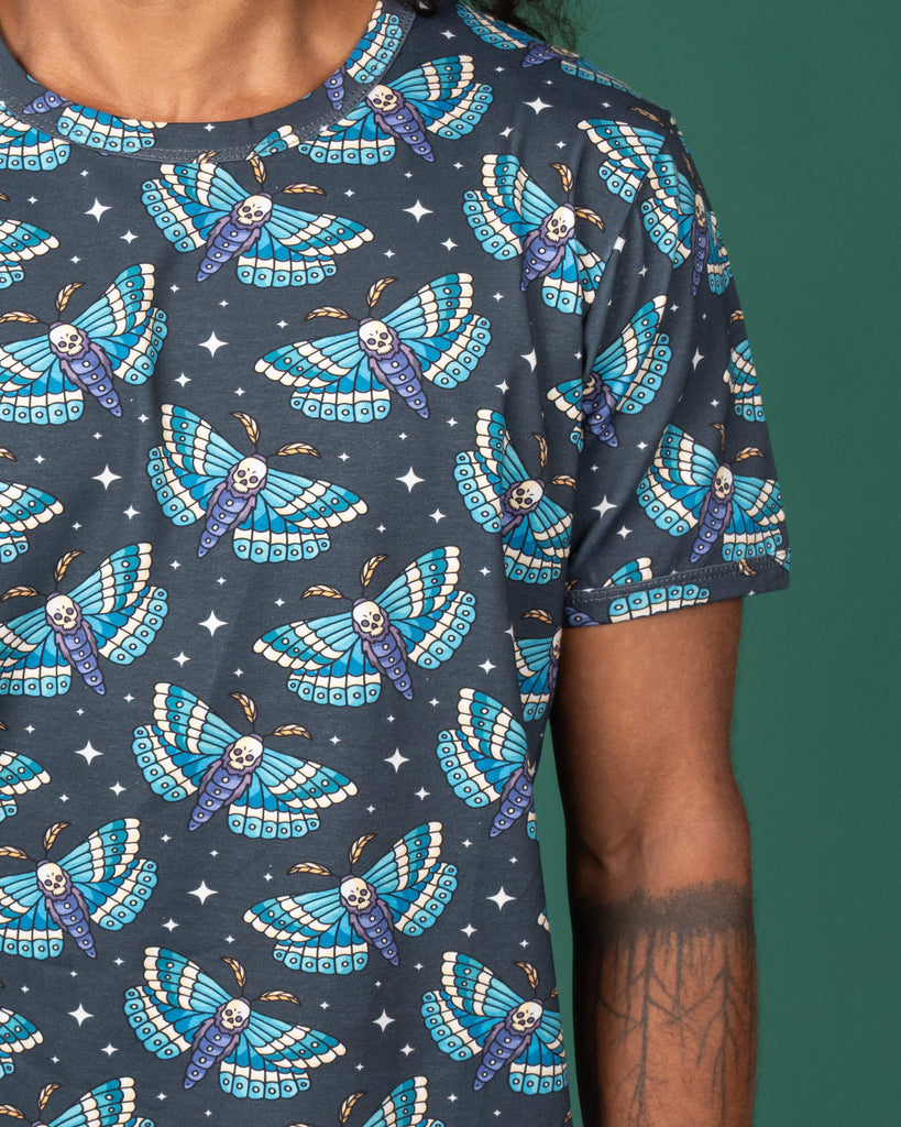 Close-up of a dark grey T-shirt from Run & Fly, featuring a colourful print of blue and teal death head moths with skulls in the centre. The model's arm is visible, displaying tattoos. The ethical slow fashion brand uses a gothic theme with bold, vibrant design choices.