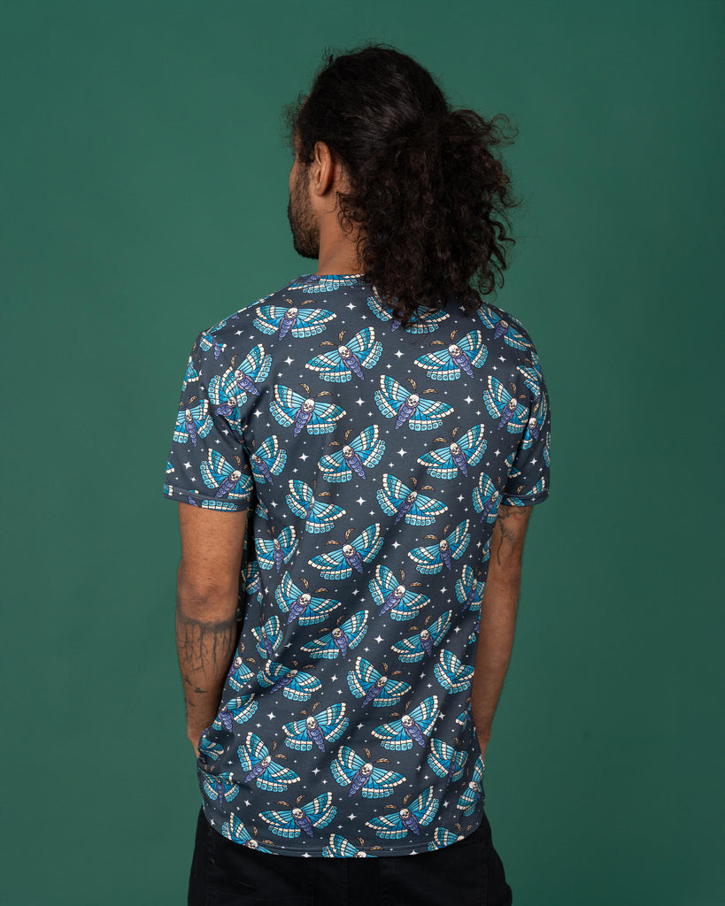 Model with dark curly hair tied back, wearing a dark grey T-shirt with a repeat print of colourful death head moths. The moths feature skulls in the centre, with vibrant blue and teal wings. The model is turned away, showing the back of the shirt, standing against a green backdrop. Ethical slow fashion by Run & Fly, highlighting alternative gothic style.