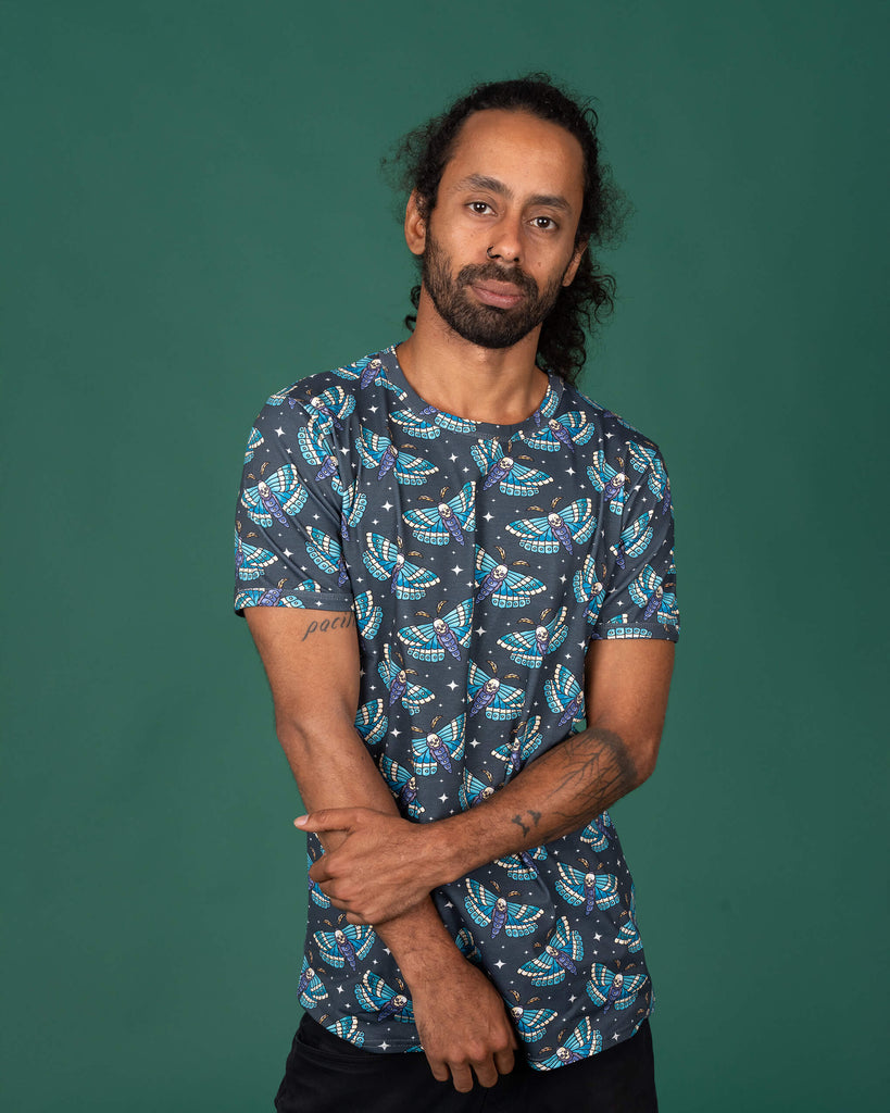 Model with dark curly hair, standing with arms crossed in a dark grey T-shirt by Run & Fly. The shirt showcases a bold death head moth print in blue and teal. The model’s tattoos are visible. Ethical slow fashion brand, known for unique alternative designs.