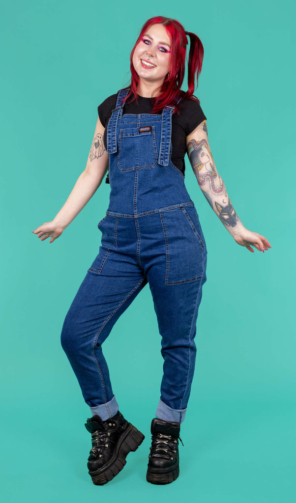 Flo a red haired femme white model with tattoos is wearing blue denim dungarees with a black tee and black platform trainers shot in a studio by Amy Davies Photography