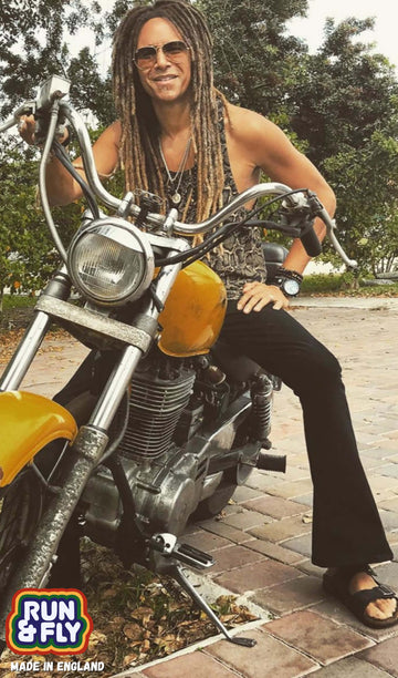Model is sat on a gold and black vintage motorbike wearing the black stretch cotton bell bottom super flares jeans with a crocodile print vest and black sandals. They are facing forward posing with one hand on the bike and the other hand resting on their leg whilst smiling to camera.