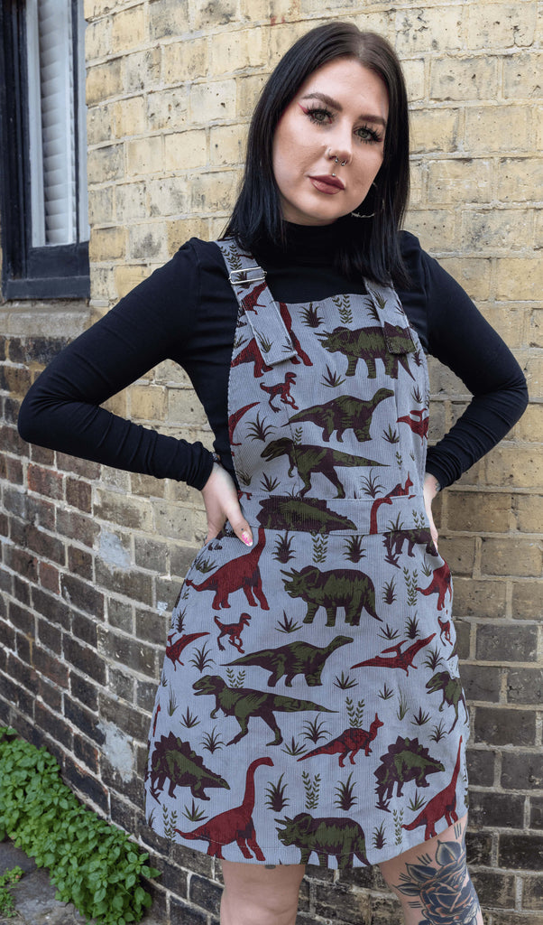 Florence is stood in front of a brick wall wearing the corduroy adventure dinosaur pinafore dress with a black long sleeve high neck top underneath. She is looking into the camera with both hands on her hips. Photo has been cropped from the knees up.