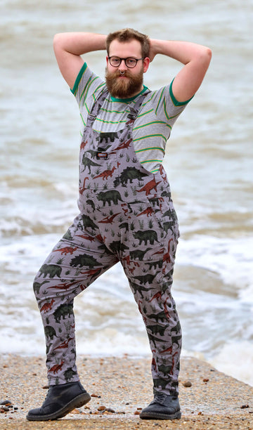 Thomas with a beard, glasses and nose ring is  smiling by the West Pier in Brighton wearing grey dinosaur twill dungarees with a striped t-shirt underneath and black boots. He is facing the camera and is posing with both hands behind his head and his left leg bent out the side.