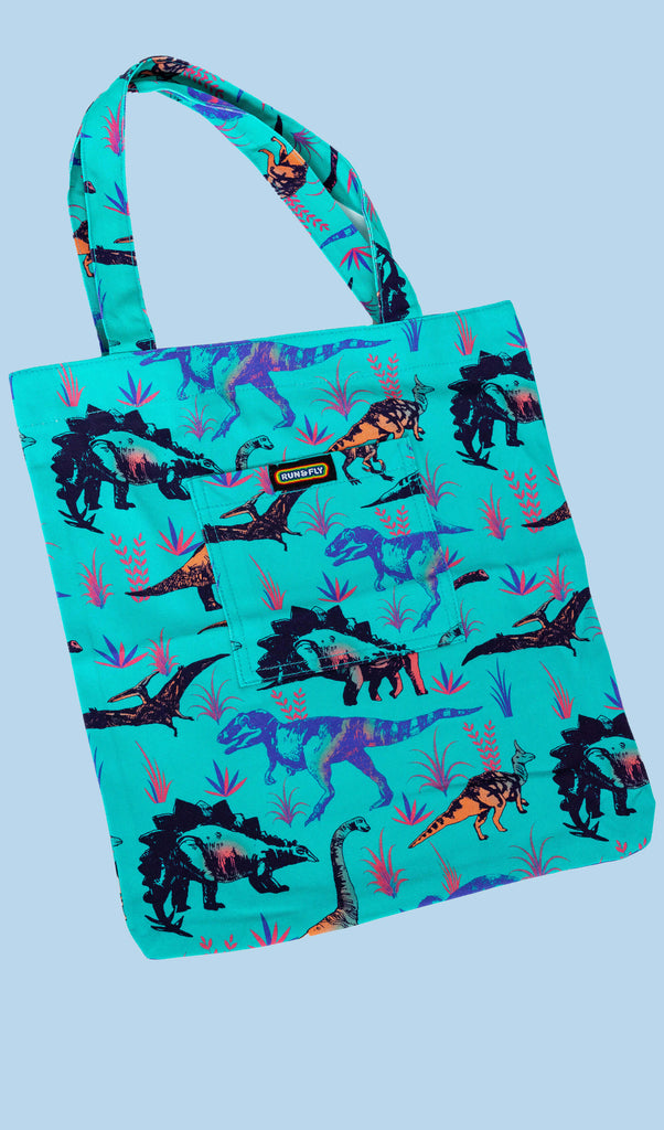 Flat lay of the Jade Adventure Dinosaur Tote Bag on a light blue backdrop. The green tote bag has an all over dinosaur print with a front pocket with rainbow Run&Fly logo on the front.