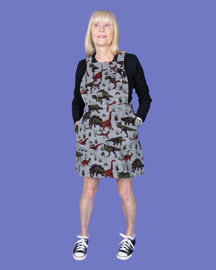 A full-length image of a woman with long blonde hair wearing a grey dinosaur print pinafore dress over a black long-sleeved top. She is standing with her hands in the pockets of the dress, which is made by Run & Fly, an ethical slow fashion company. She completes the look with black and white Converse trainers. The backdrop is a solid lavender purple.