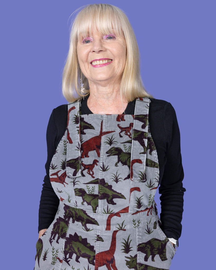 A close-up portrait of a smiling woman with long blonde hair, wearing a grey corduroy pinafore dress with a dinosaur print over a black long-sleeved top. The dress is designed by Run & Fly, a small ethical slow fashion brand. The background is a solid lavender purple.