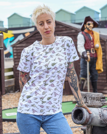 Yo is stood in a mini golf course area wearing the white jurassic dinosaur ringer print tshirt with blue denim jeans. They are facing forward posing with one hand resting on the golf course and the other behind them. Photo is cropped from the hips up.