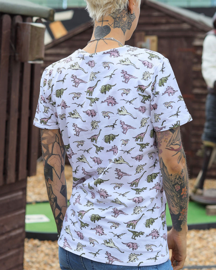 Yo is standing looking away from the camera to show the back print of the white jurassic dinosaur ringer t-shirt. They have many tattoos on their arms and hands and in their front pockets.