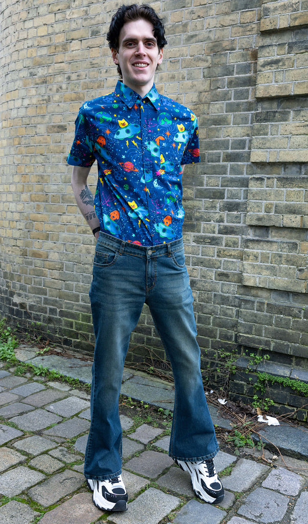 Jake a man with dark hair and tattoos is wearing Unisex Distressed Denim Bellbottom Flares Mid Rise Jeans paired with Dogs in Space shirt and white trainers. Jake is smiling in front of a brick wall with his arms behind him and arms behind his back.
