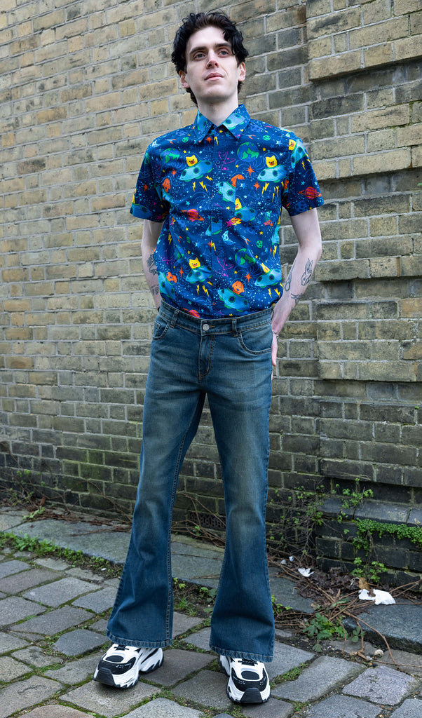 Jake a man with dark hair and tattoos is wearing Unisex Distressed Denim Bellbottom Flares Mid Rise Jeans paired with Dogs in Space shirt and white trainers. Jake is stood in front of a brick wall outside and is looking into the distance with his hands on his hips.