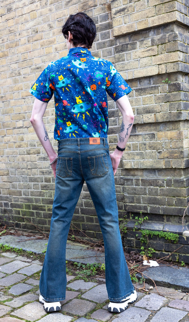 Jake a man with dark hair and tattoos is wearing Unisex Distressed Denim Bellbottom Flares Mid Rise Jeans paired with Dogs in Space shirt and white trainers.  Jake is stood with his back to the camera in front of a brick wall with his arms out by his sides. 