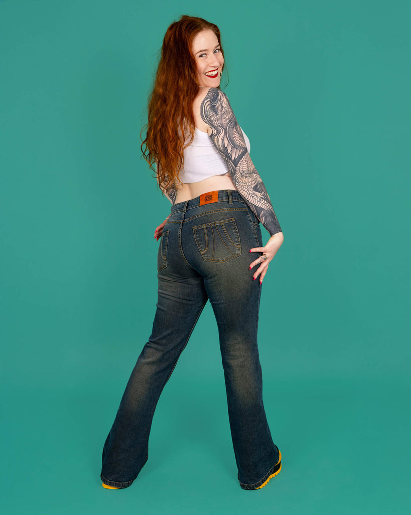 Isobella a tattooed model with long ginger hair is wearing Unisex Distressed Denim Bellbottom Flares Mid Rise Jeans paired with a white crop top and black and yellow boots. The model is stood with her back to the camera and looking bac over her shoulder and smiling at the camera. The background of the photo is green.