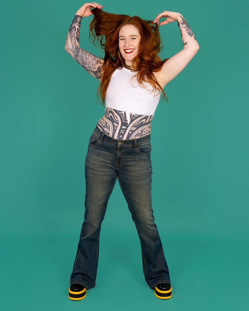 Isobella a tattooed model with long ginger hair is wearing Unisex Distressed Denim Bellbottom Flares Mid Rise Jeans paired with a white crop top and black and yellow boots. She is smiling with her hands in her hair in front of a green background.