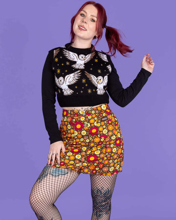 Florence is wearing Ditsy Floral A Line Skirt paired with a black jumper, fishnet tights and black boots. The skirt is a dark green colour and is covered in red, orange and yellow flowers. The model is posing with one leg bent and flicking her hair. The background of the photo is purple.