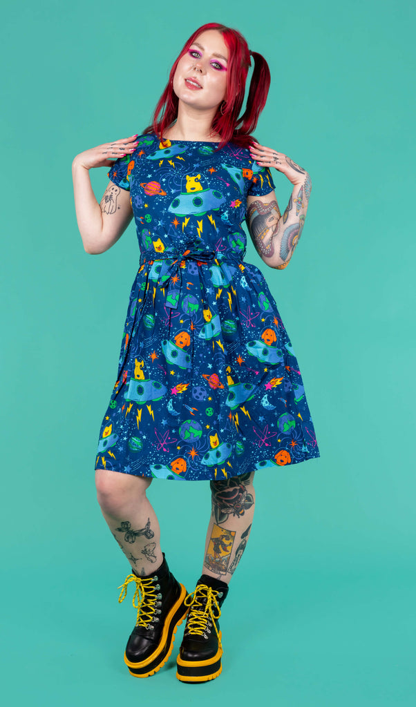 The Dogs in Space Stretch Belted Tea Dress with Pockets worn by Flo on a teal background. Flo has red hair in bunches, neon pink makeup and tattoos with yellow and black platform boots. The print features orange and yellow bull terriers, pomeranians and labradors in blue spaceships on a space themed blue background with stars, planets and rockets. The dress is short sleeved with pockets and a belt tie. She is facing forward smiling with both hands resting on her shoulders.