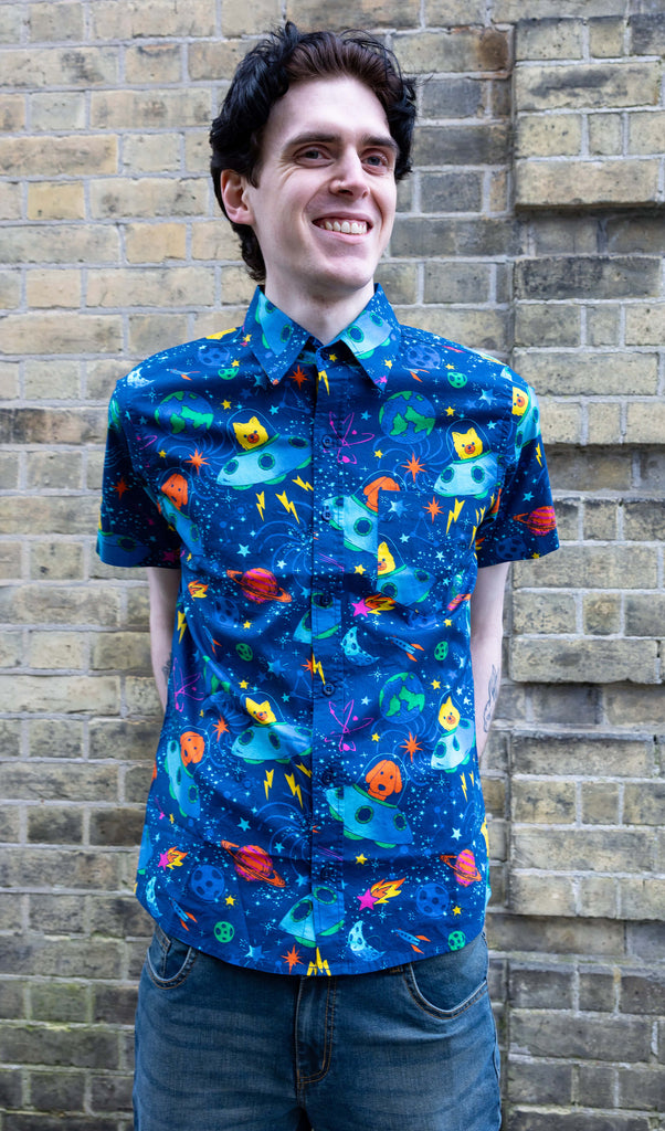 Jake a white male with dark hair is wearing Dogs in Space Short Sleeve Shirt paired with blue jeans. The bright blue shirt as yellow and orange dogs in little spaceships with various rocket, stars and planets all over. Jake is smiling and looking into the distance with his hands behind his back in front of a brick wall in Hove. 