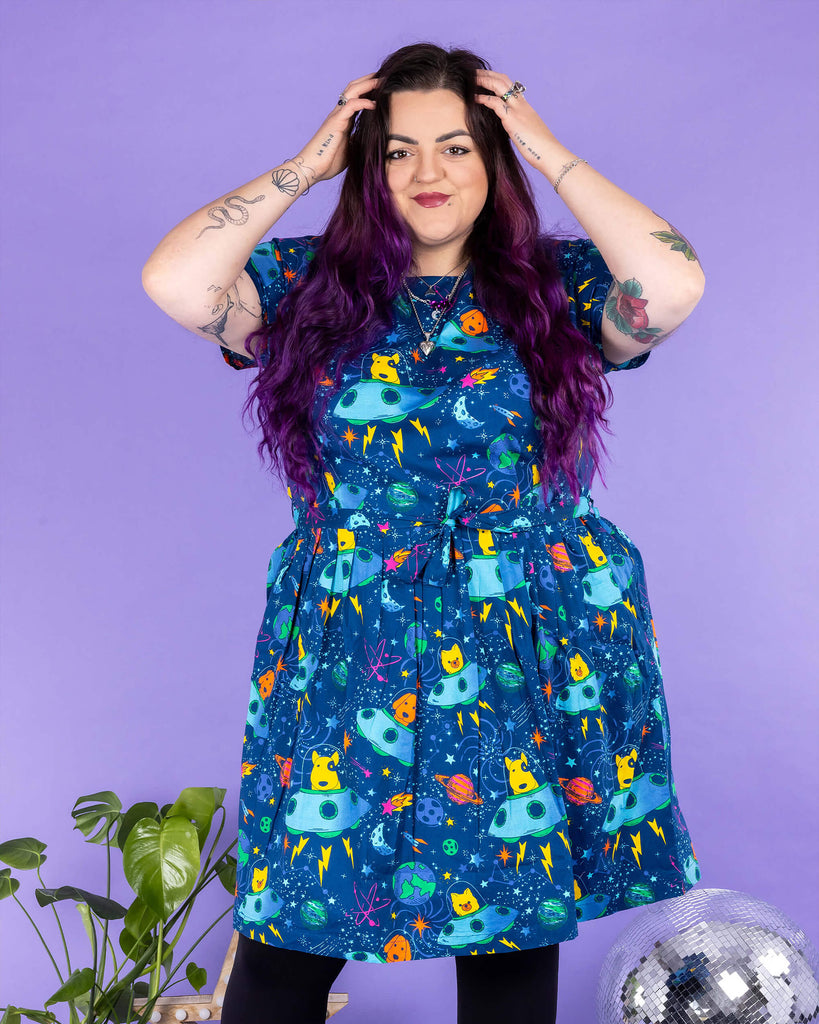 Luisa, a femme model with purple hair and tattoos, is stood in a photography studio in Hove in front of a purple backdrop wearing Dogs in Space Stretch Belted Tea Dress with Pockets with black tights and boots. The print features orange and yellow bull terriers, pomeranians and labradors in blue spaceships on a space themed blue background with stars, planets and rockets. The dress is short sleeved with pockets and a belt tie. Luisa is facing the camera posing with her hands in her hair.