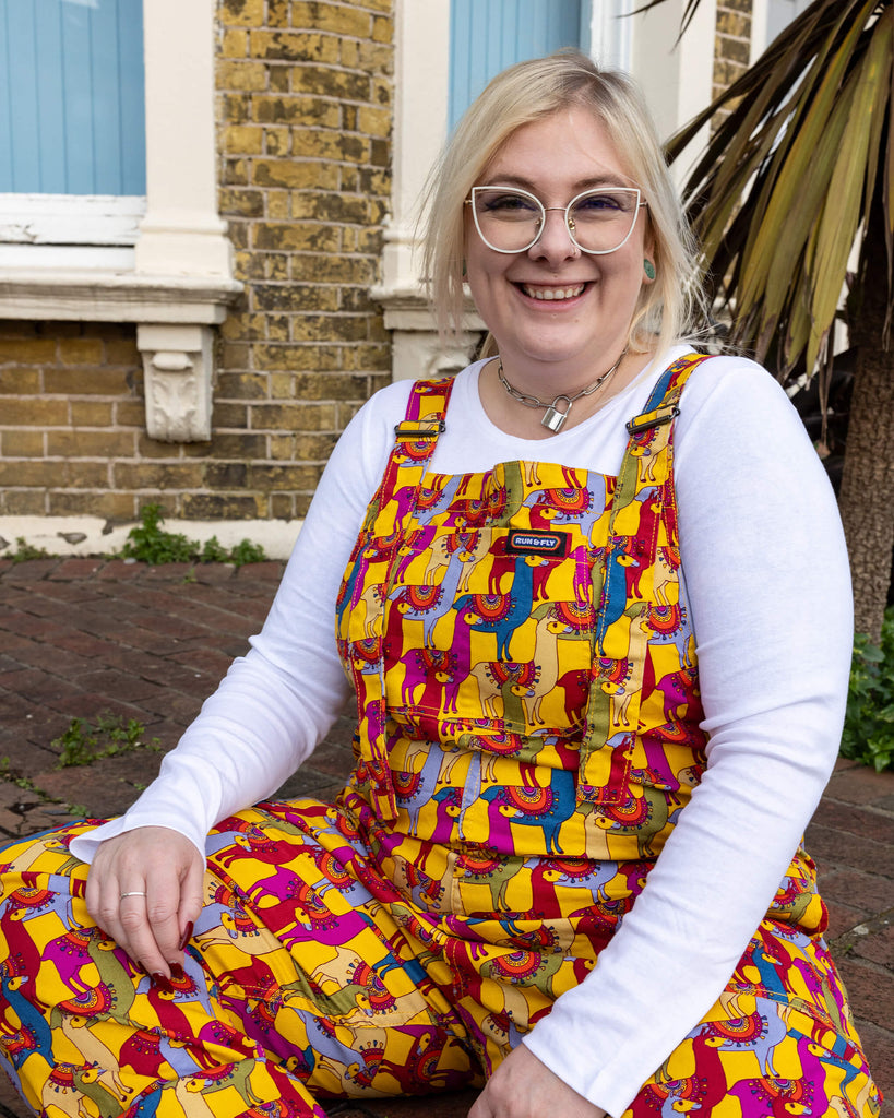 Close-up of a person wearing Run & Fly drama llama dungarees, seated in an outdoor setting. The dungarees showcase a bold, playful llama print with bright colours, including yellow, pink, and blue. Paired with a white top and accessorised with a silver necklace. Run & Fly is a UK-based ethical slow fashion brand focused on unique, eco-friendly clothing.