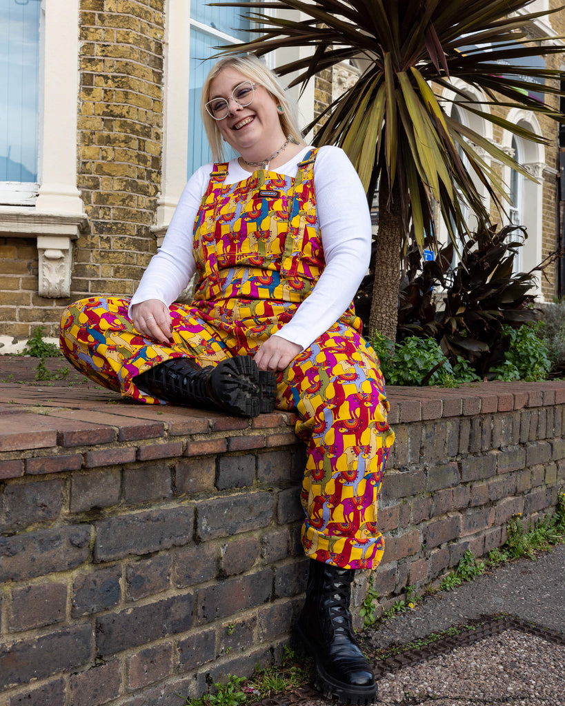 A smiling person sitting on a brick ledge, wearing Run & Fly drama llama dungarees. The dungarees feature a vibrant print of colourful llamas in shades of yellow, pink, and blue with intricate detailing. Paired with black combat boots and a white long-sleeved top. Run & Fly is a small ethical slow fashion brand known for quirky and sustainable designs.
