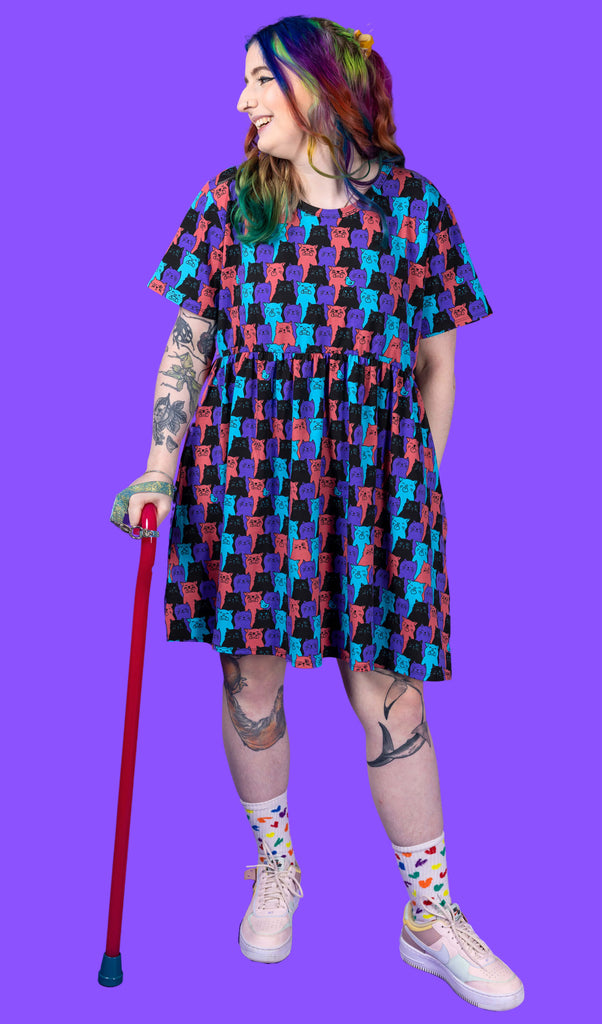 Non-binary disabled femme model with rainbow coloured hair is wearing Cat Chorus Oversized Smock Dress, paired with rainbow coloured socks and pink trainers. The dress has an all over print of purple, black, red and blue cats.
